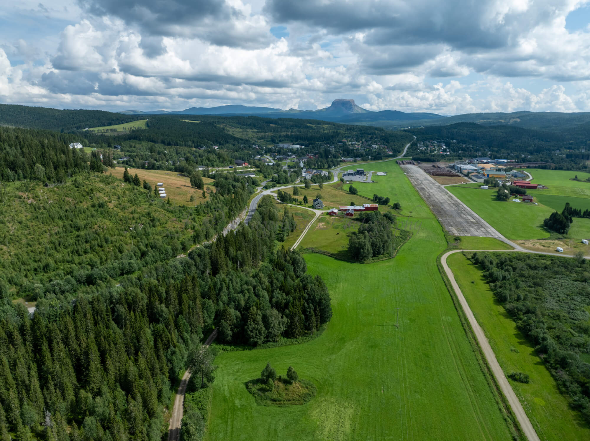 An old airstrip  and fields with buldings beyond it and mountains on the background where one looks like a hat
