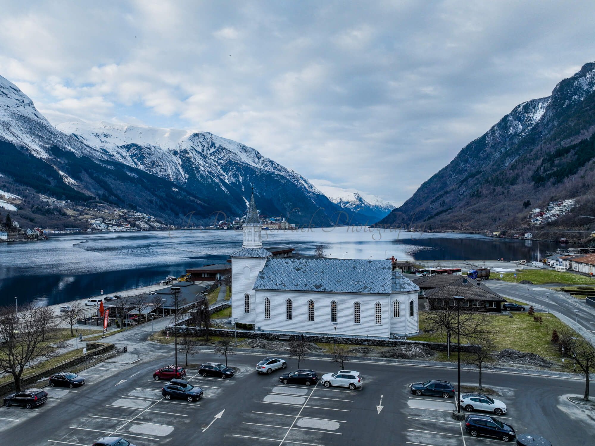 A white church in a town with a parking lot in front and a large body of water behind with land on both sides